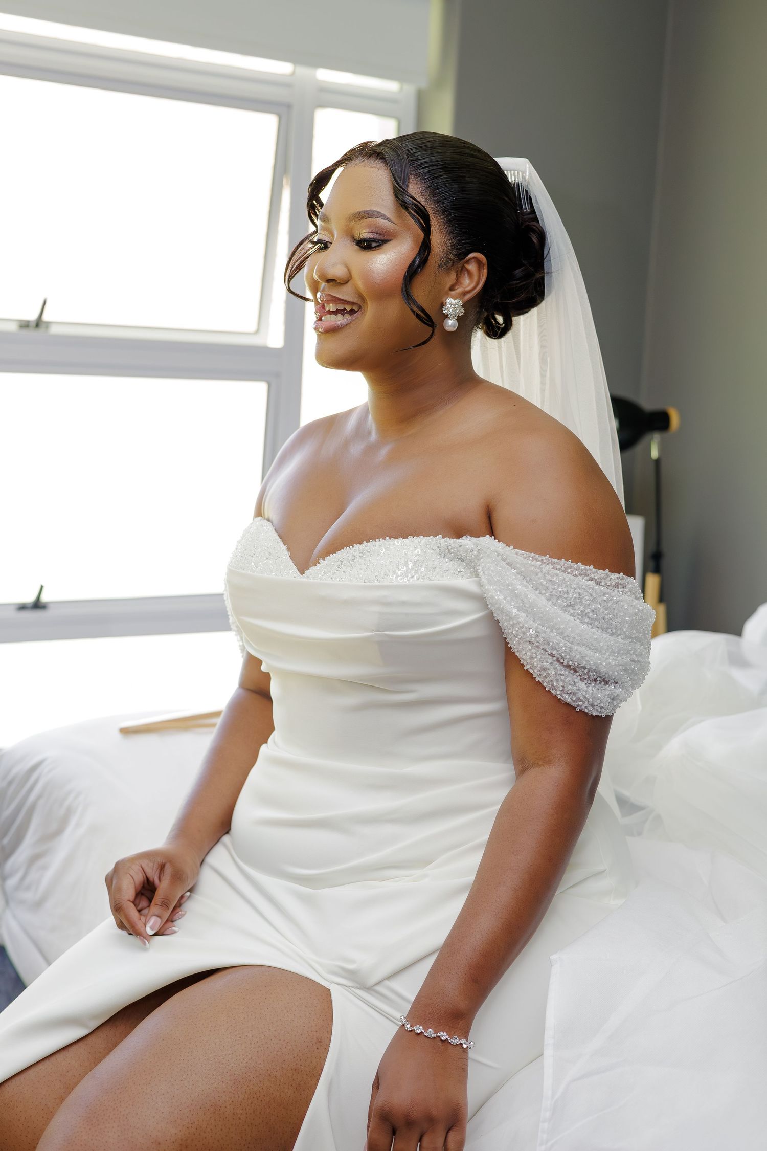 Radiant bride in off-shoulder white wedding gown with veil and pearl earrings sitting by window.