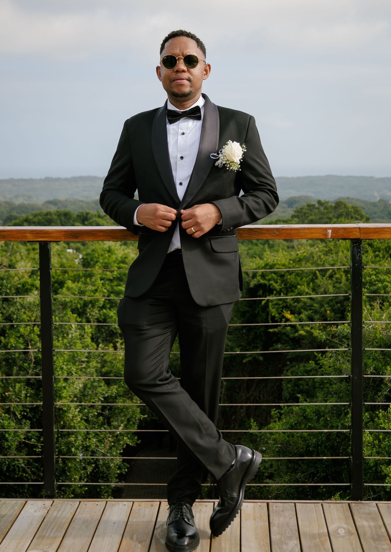 Stylish groom in black tuxedo with boutonniere and sunglasses posing on a wooden deck outdoors.