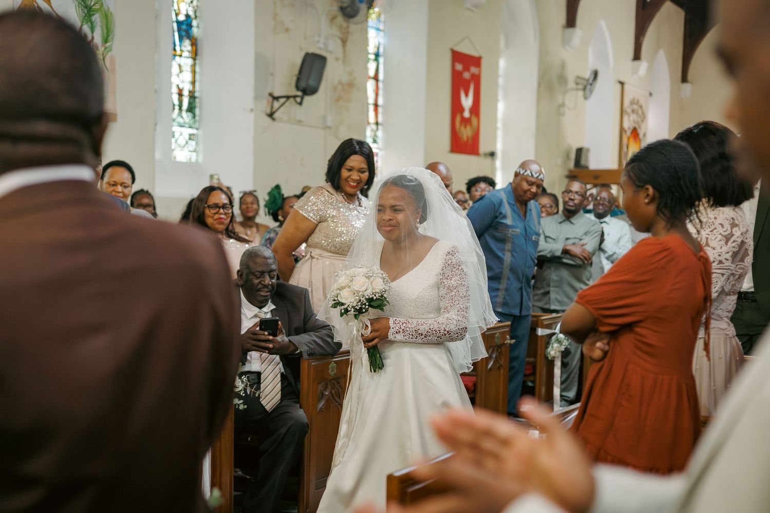Bride walks down church aisle in white lace gown, smiling at guests during wedding ceremony.