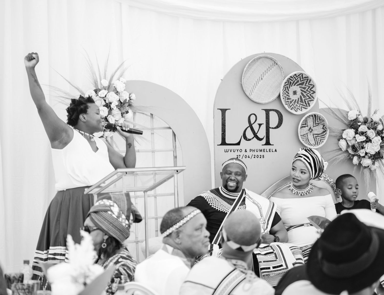 Black and white photo of a woman performing at a traditional African wedding ceremony with L&P signage backdrop.