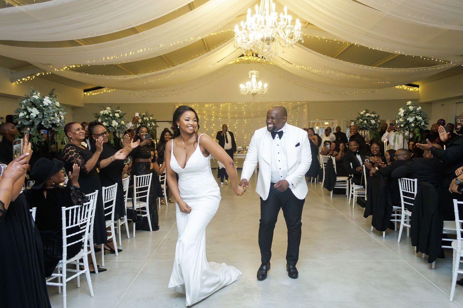 Joyful bride and groom enter their elegant wedding reception hall to cheers from guests in black attire.