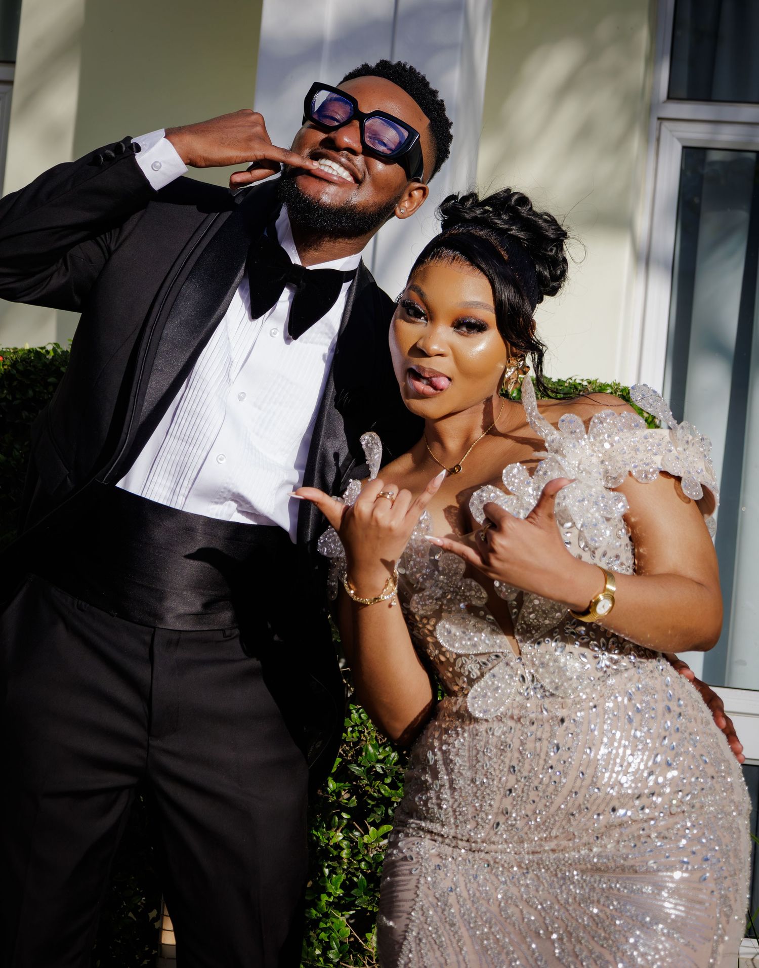 Couple posing playfully outdoors, man in black tuxedo and woman in silver beaded gown.