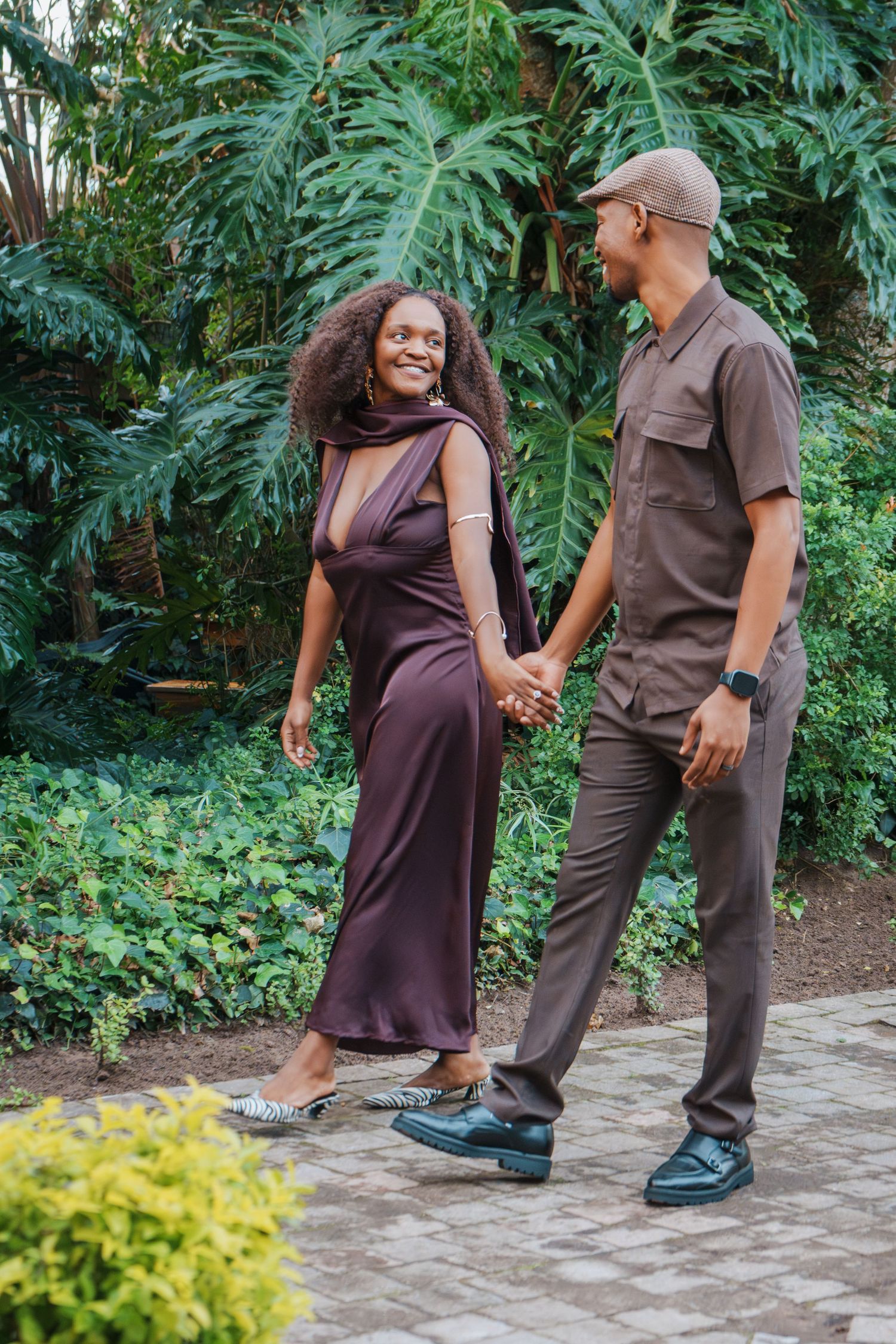 A couple in matching brown outfits holds hands while walking through a lush tropical garden, smiling at each other.
