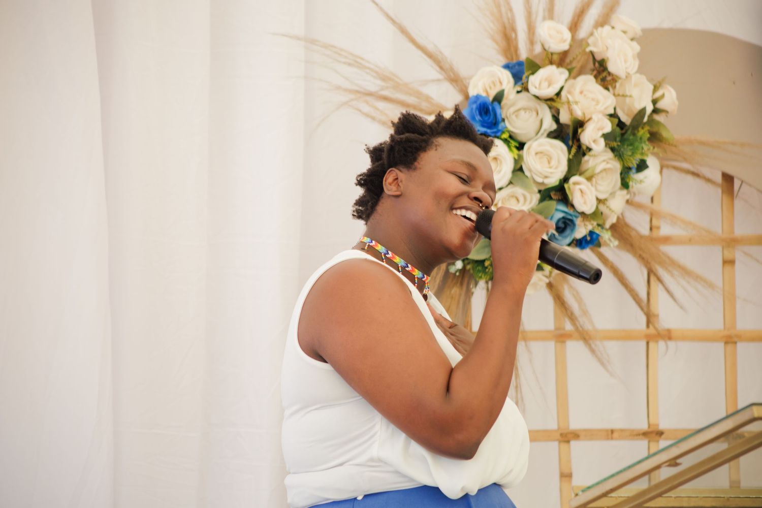 Woman singing joyfully into a microphone at a wedding reception with white and blue floral backdrop.