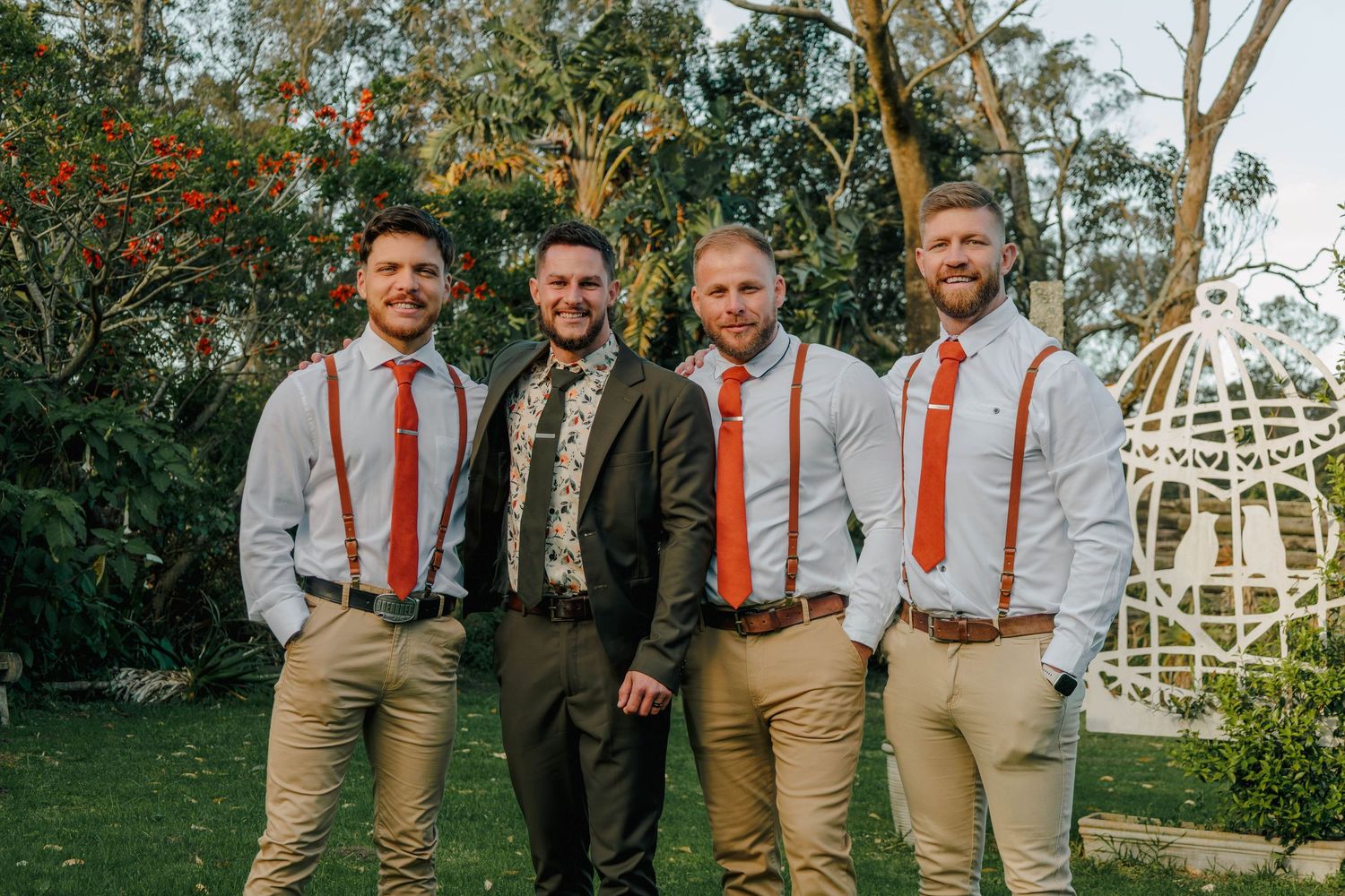 Groom poses with three groomsmen wearing orange ties and suspenders in sunny garden setting.