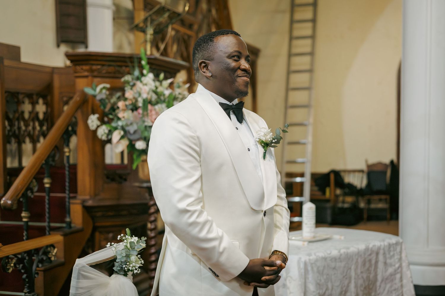 Happy groom in white tuxedo with boutonniere smiles at church altar.
