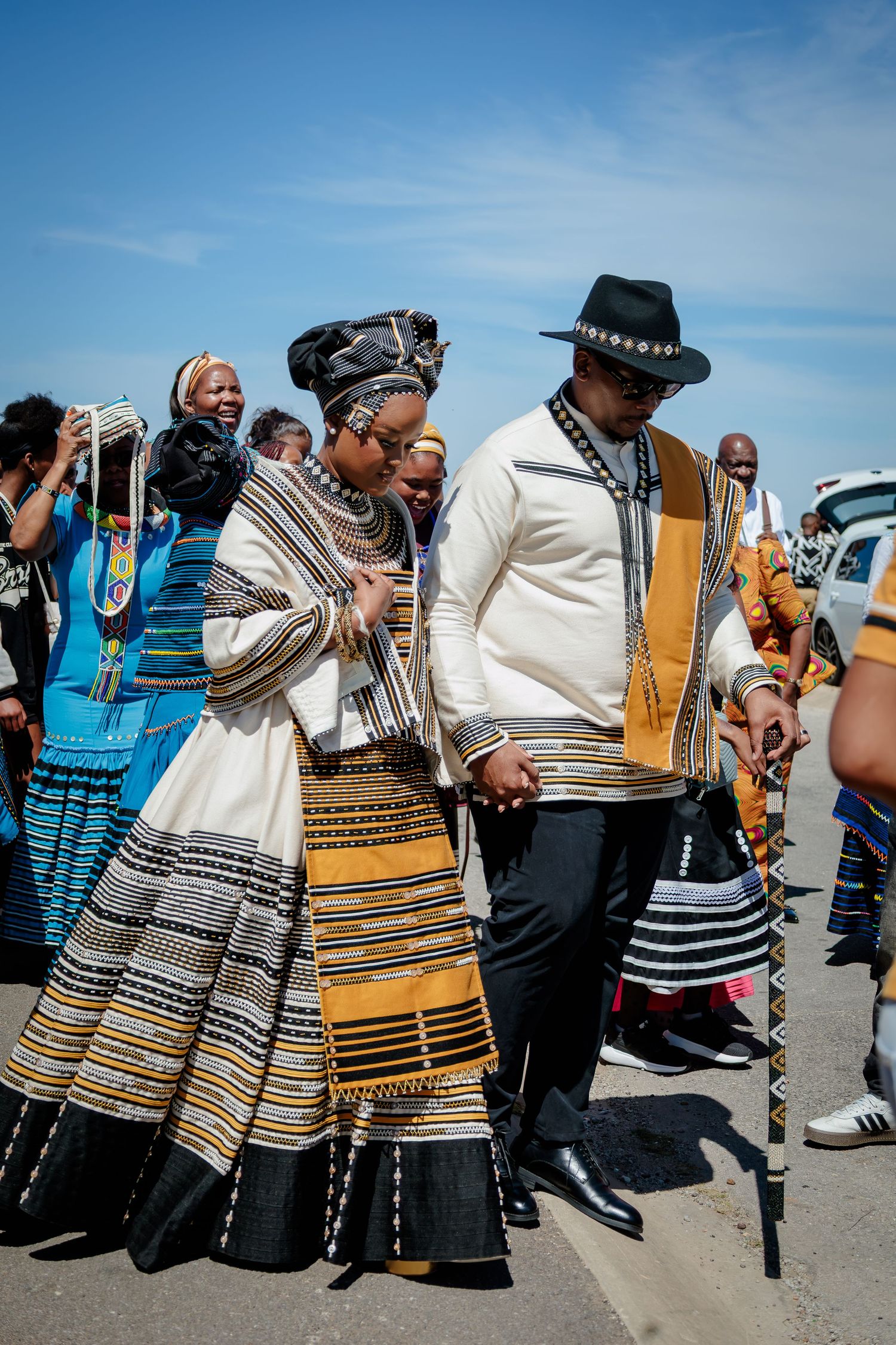 Couple in matching traditional Xhosa wedding attire walk hand in hand during an outdoor ceremony.