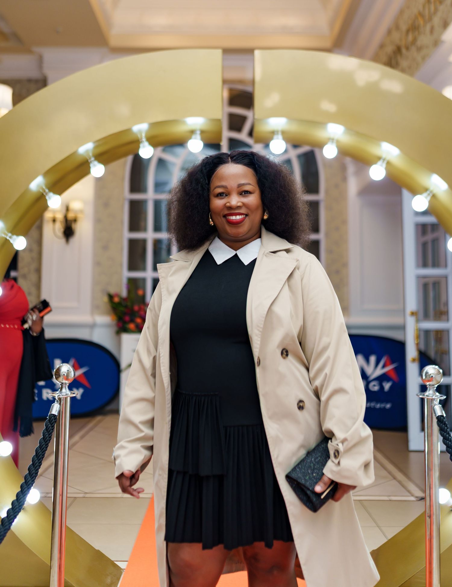 Woman in beige trench coat and black dress posing at Beyond Energy corporate event.