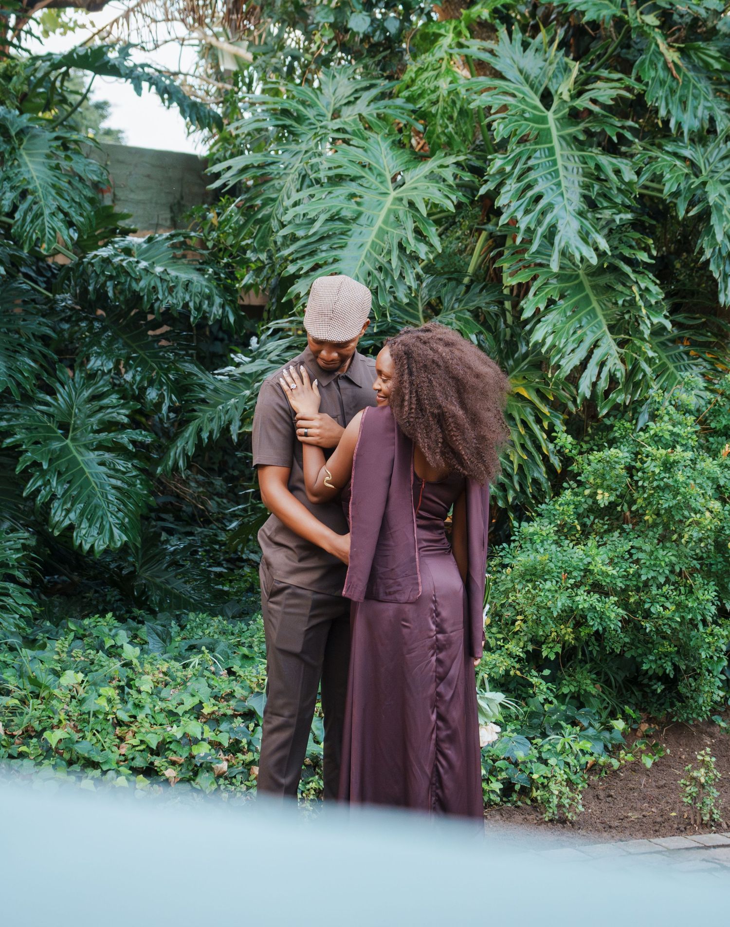A couple in elegant brown attire embraces tenderly among lush tropical greenery in an outdoor setting.