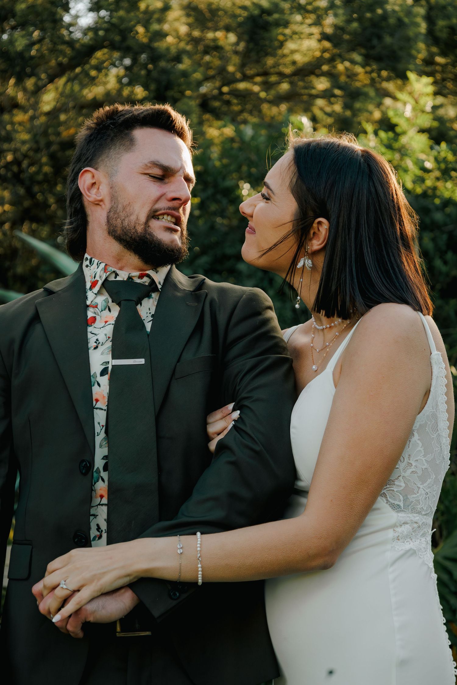 Bride and groom share a playful intimate moment outdoors, with groom in dark suit and floral tie.