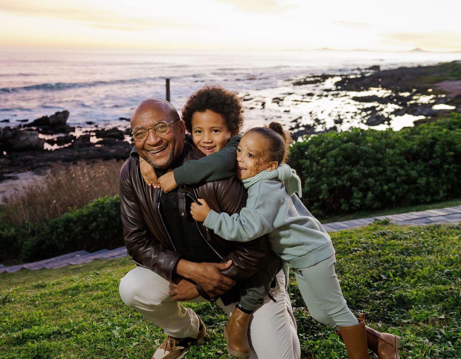Joyful grandfather kneeling on grass hugging two young grandchildren with ocean backdrop at sunset.