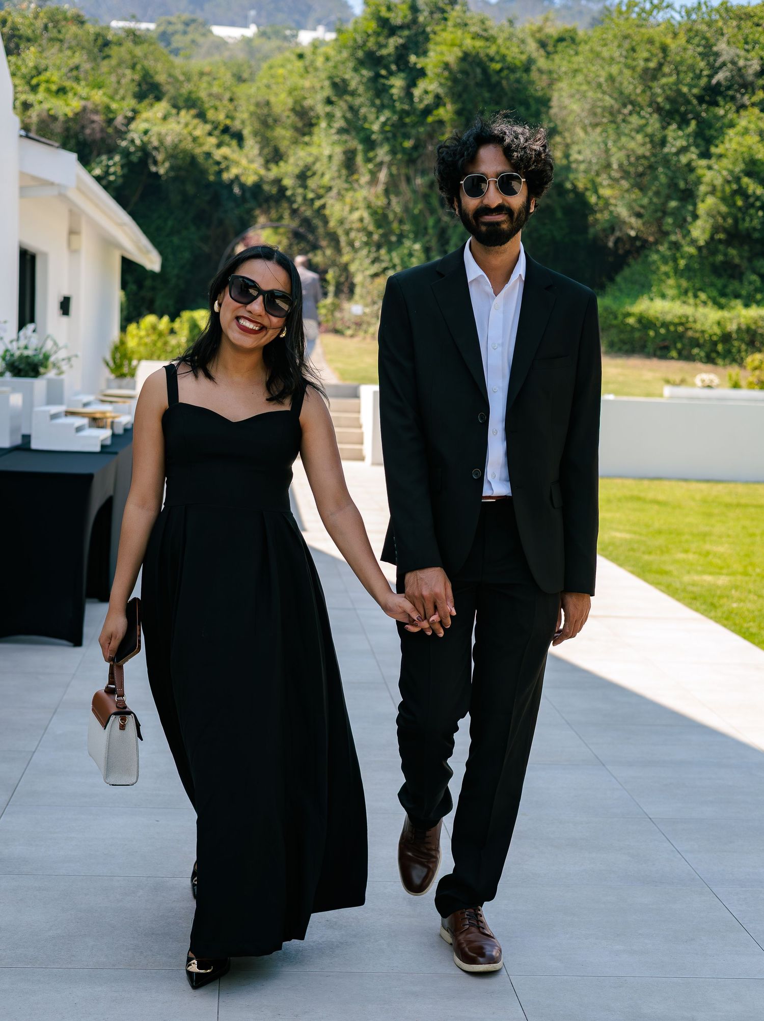 Elegant couple in matching black outfits holding hands and walking on a patio at an outdoor wedding venue.