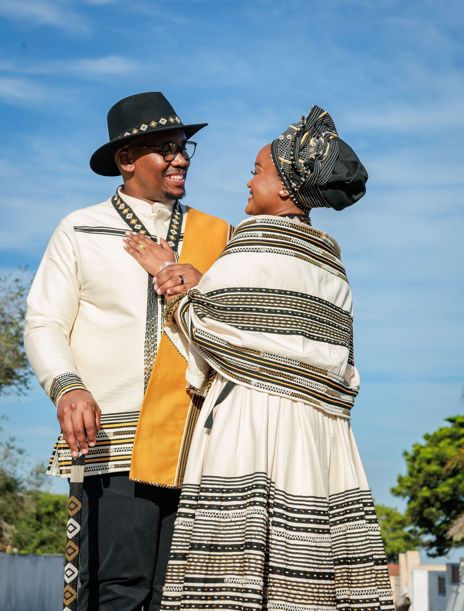 Couple in traditional Xhosa attire, man in white outfit with black hat, woman in striped dress with head wrap.