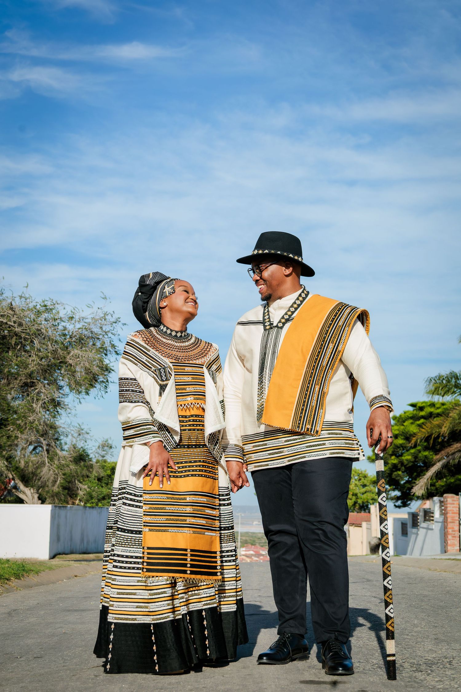Couple in traditional Xhosa ceremonial dress holding hands and gazing at each other on a sunny street.