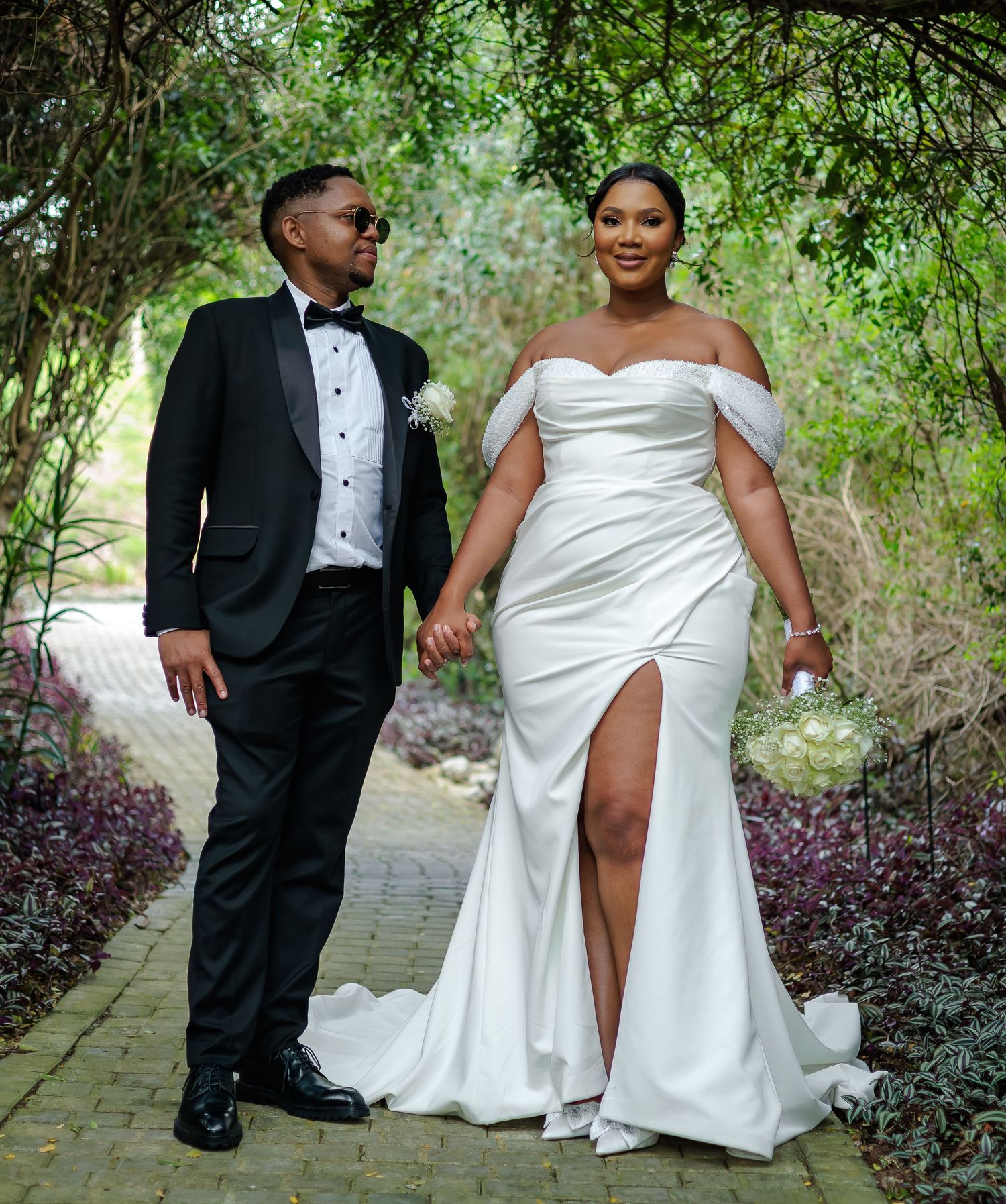 Elegant bride and groom in tuxedo pose hand-in-hand on garden path at wedding.
