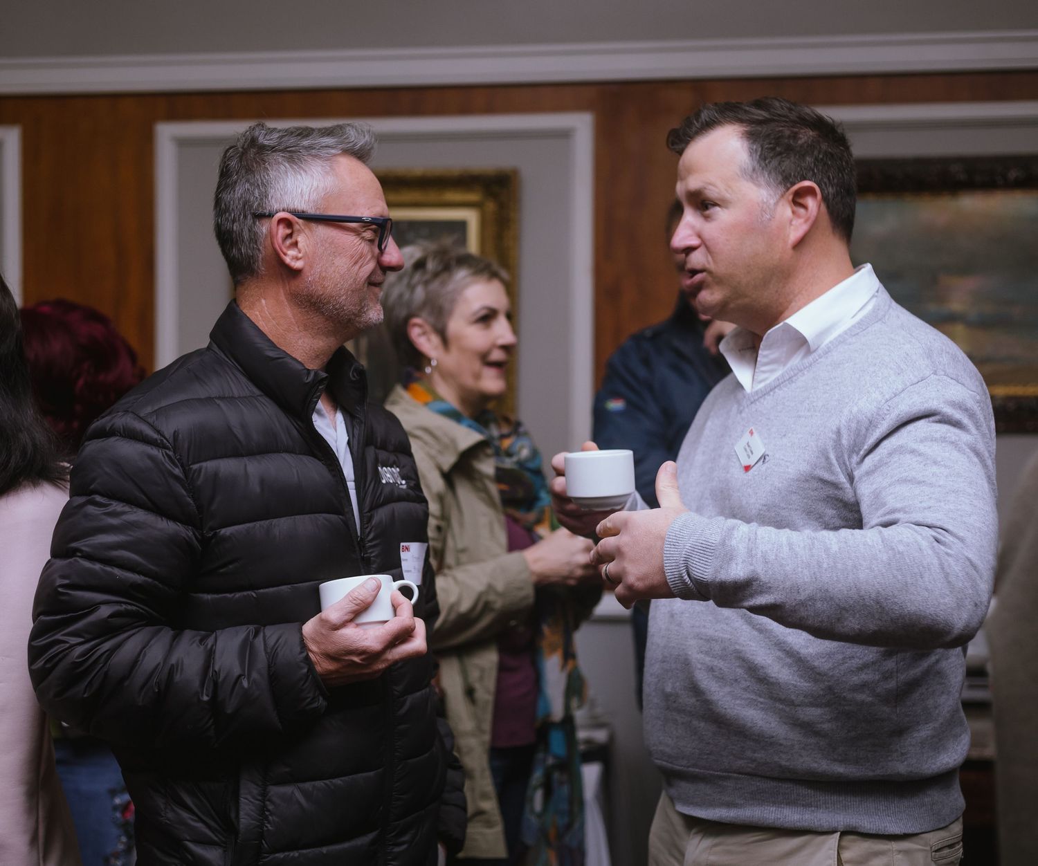 Two men holding coffee cups engaged in conversation at an indoor social gathering event.