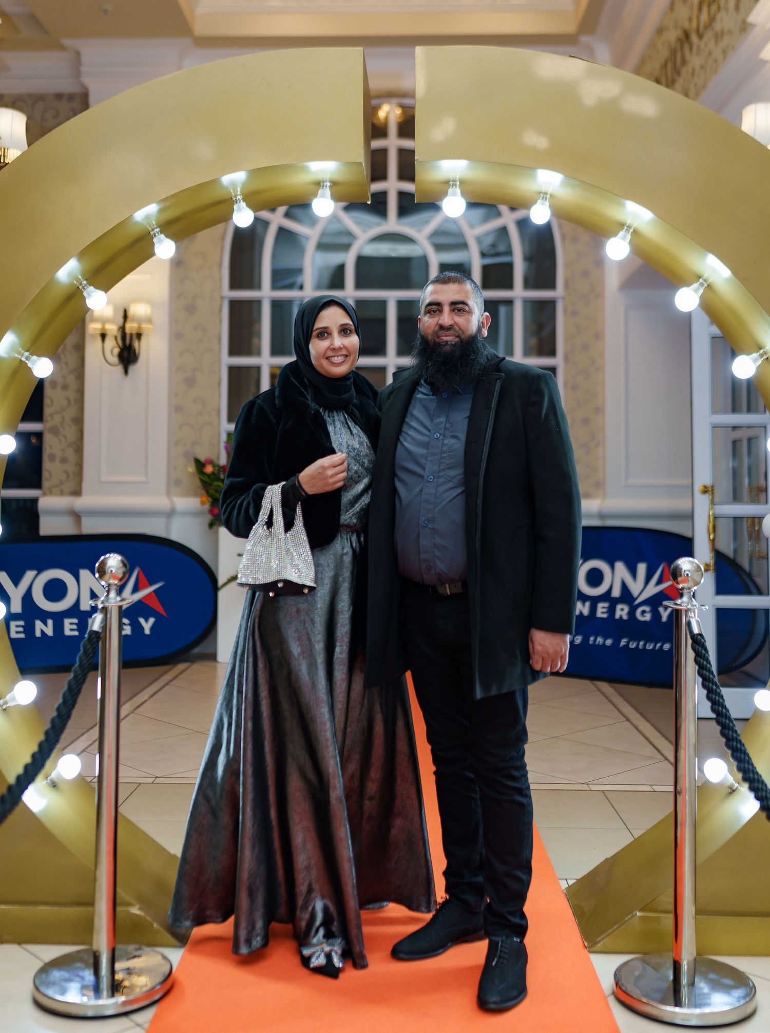 Couple posing on orange carpet at Beyond Energy event, woman in hijab and silver gown.