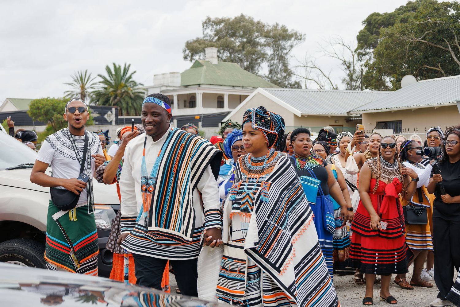 A group of people dressed in traditional Xhosa attire with striped blankets participate in a cultural ceremony.