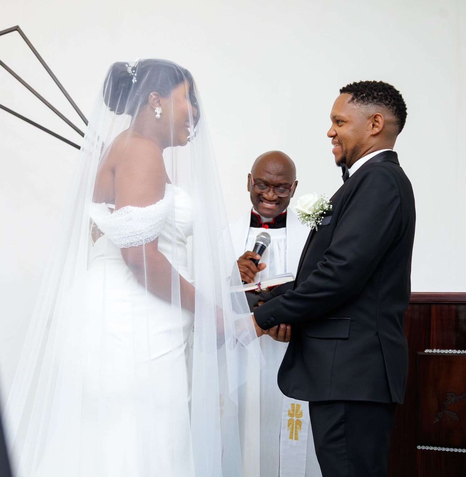 Bride and groom exchange vows at altar while officiant holds microphone in bright white chapel.