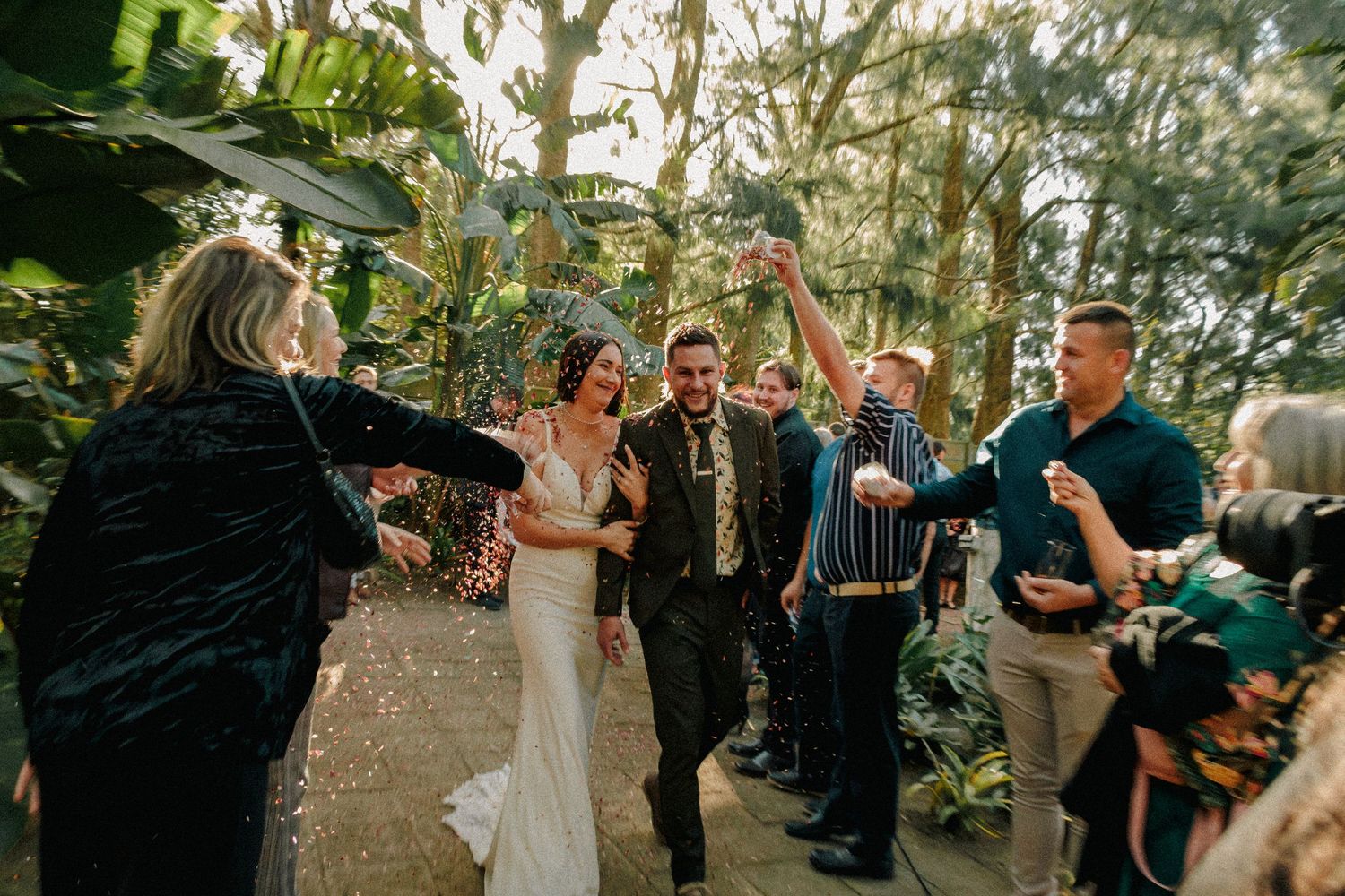 Bride and groom walk through confetti shower surrounded by cheering guests in lush garden wedding ceremony.