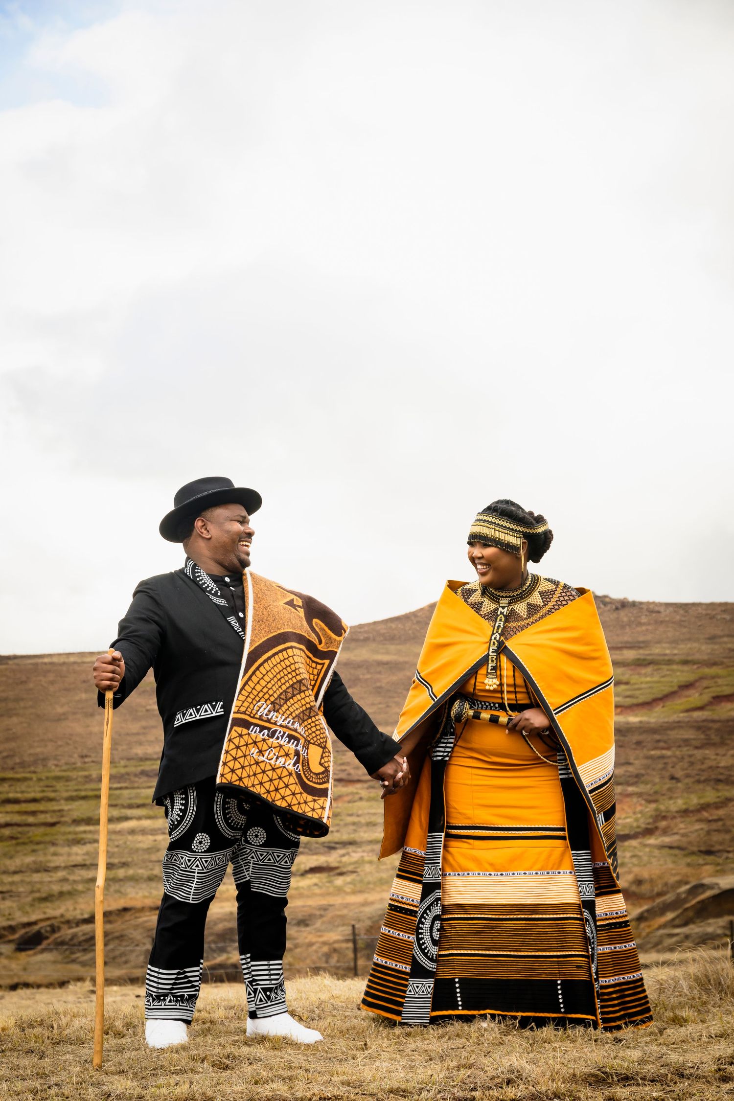 A couple in traditional African Ndebele attire holding hands in a scenic landscape, wearing vibrant orange and black patterned clothing.