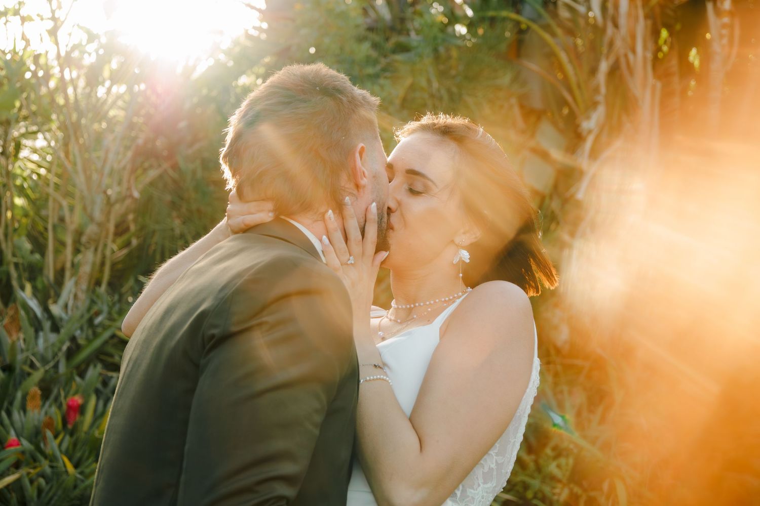 Bride and groom sharing a passionate kiss surrounded by warm golden sunlight in a garden setting.