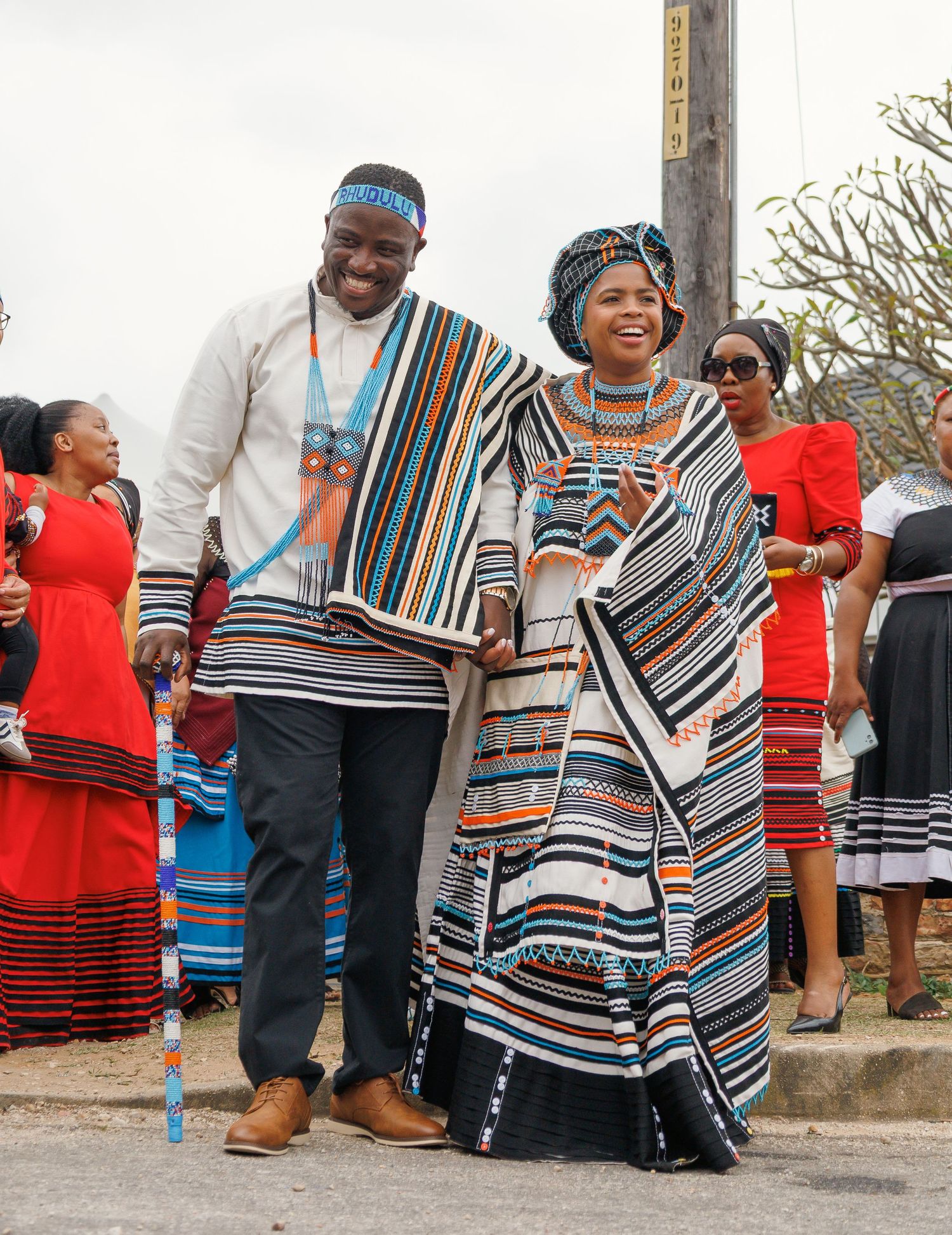 A smiling couple dressed in traditional Xhosa striped blankets and beadwork walks together at a cultural event.