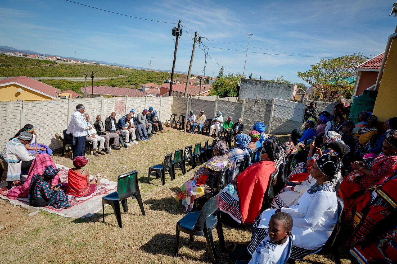 A large community gathering sits in a circle of chairs outdoors in a township setting under a bright sky.