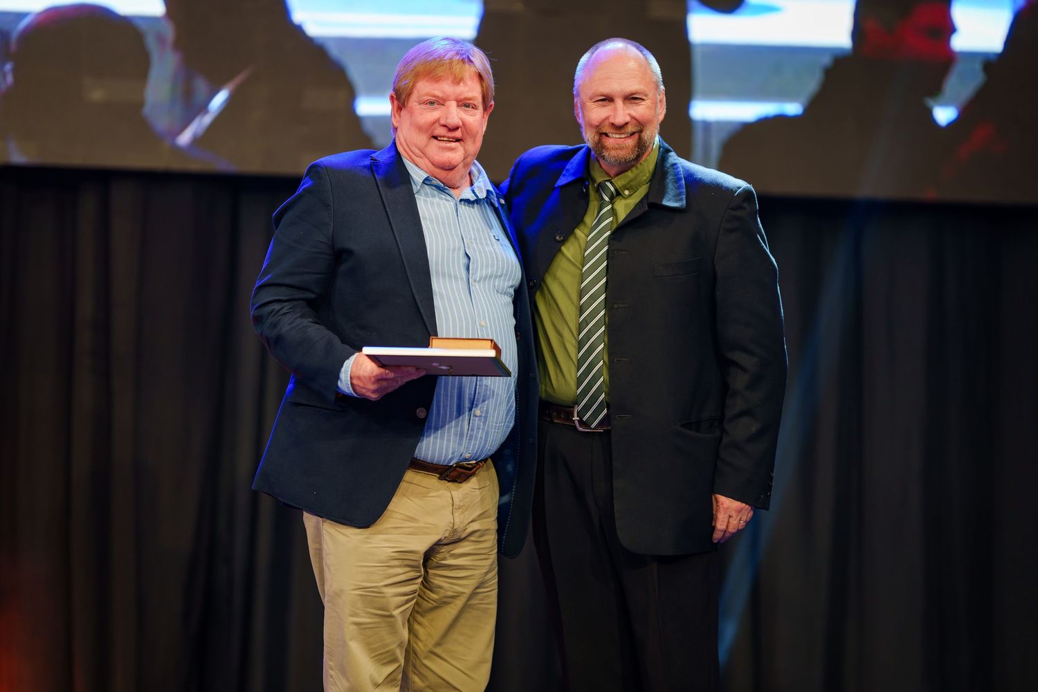 Two men pose together smiling while holding award at corporate recognition ceremony.