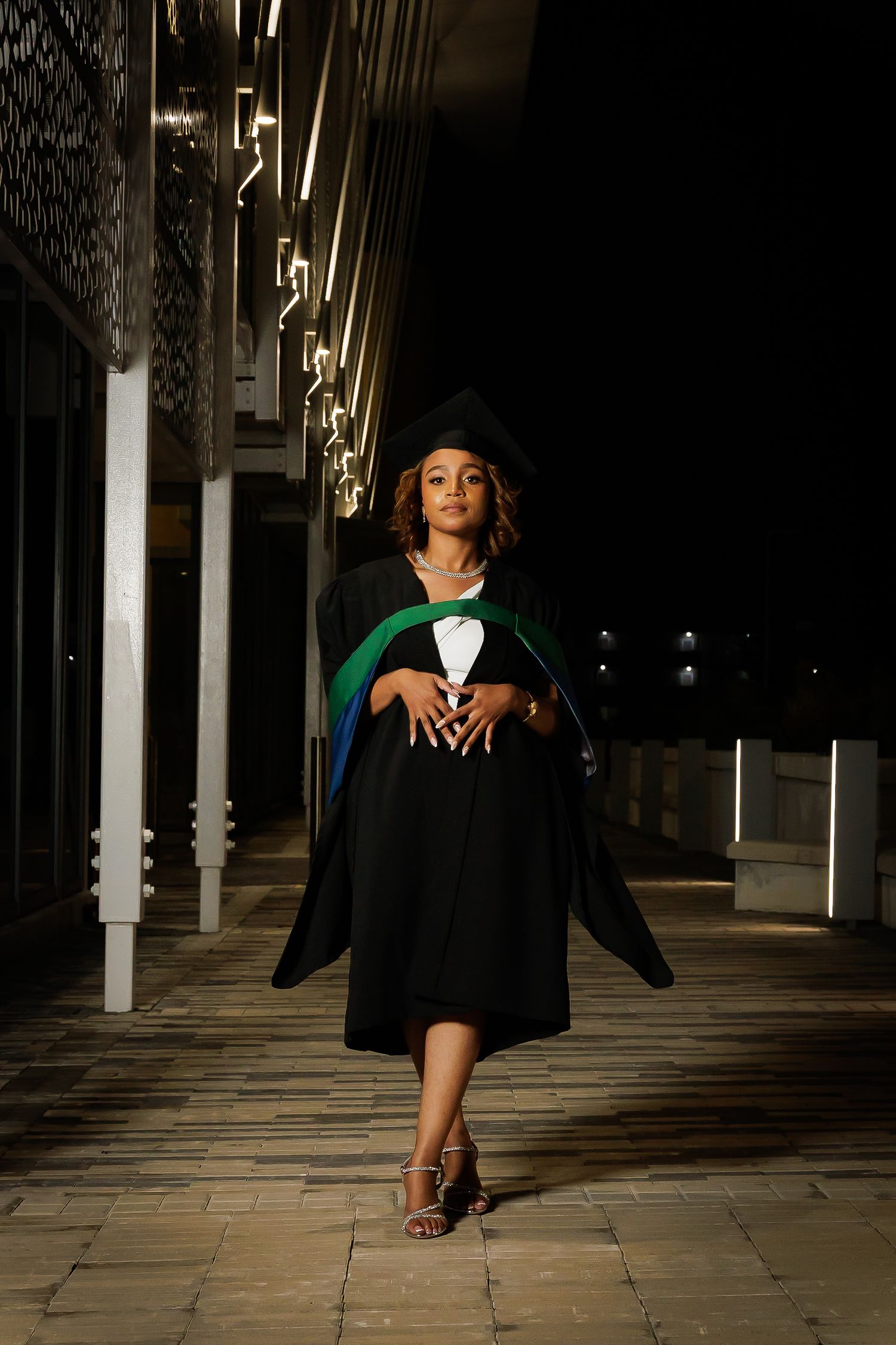 Young woman in black graduation gown and cap poses outdoors at night under lights.