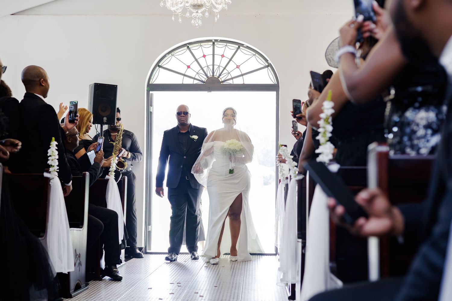 Bride in white dress walks down church aisle escorted by father toward waiting groom.