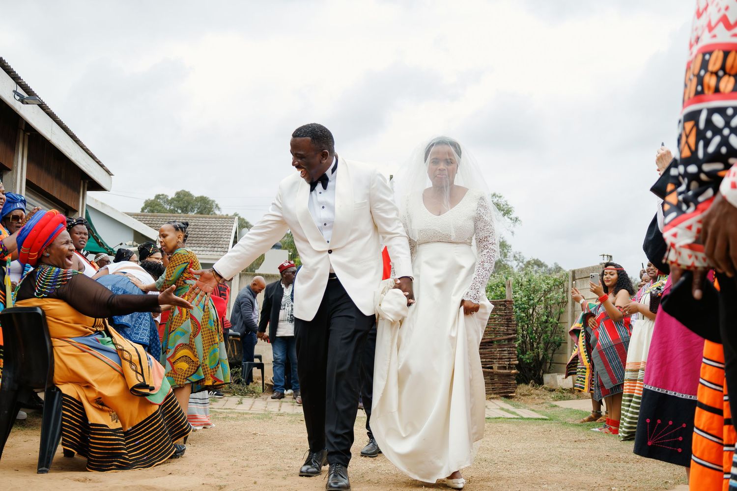 A couple in white wedding attire walks through a crowd of colorfully dressed guests at a traditional African wedding ceremony.
