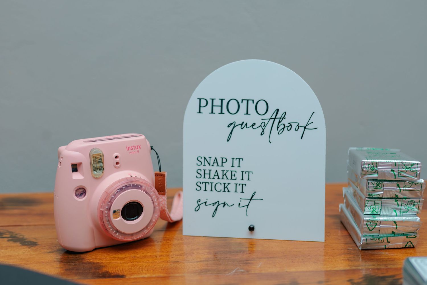 Pink Instax mini camera beside a white arch photo guestbook sign with snap, shake, stick, and sign instructions.