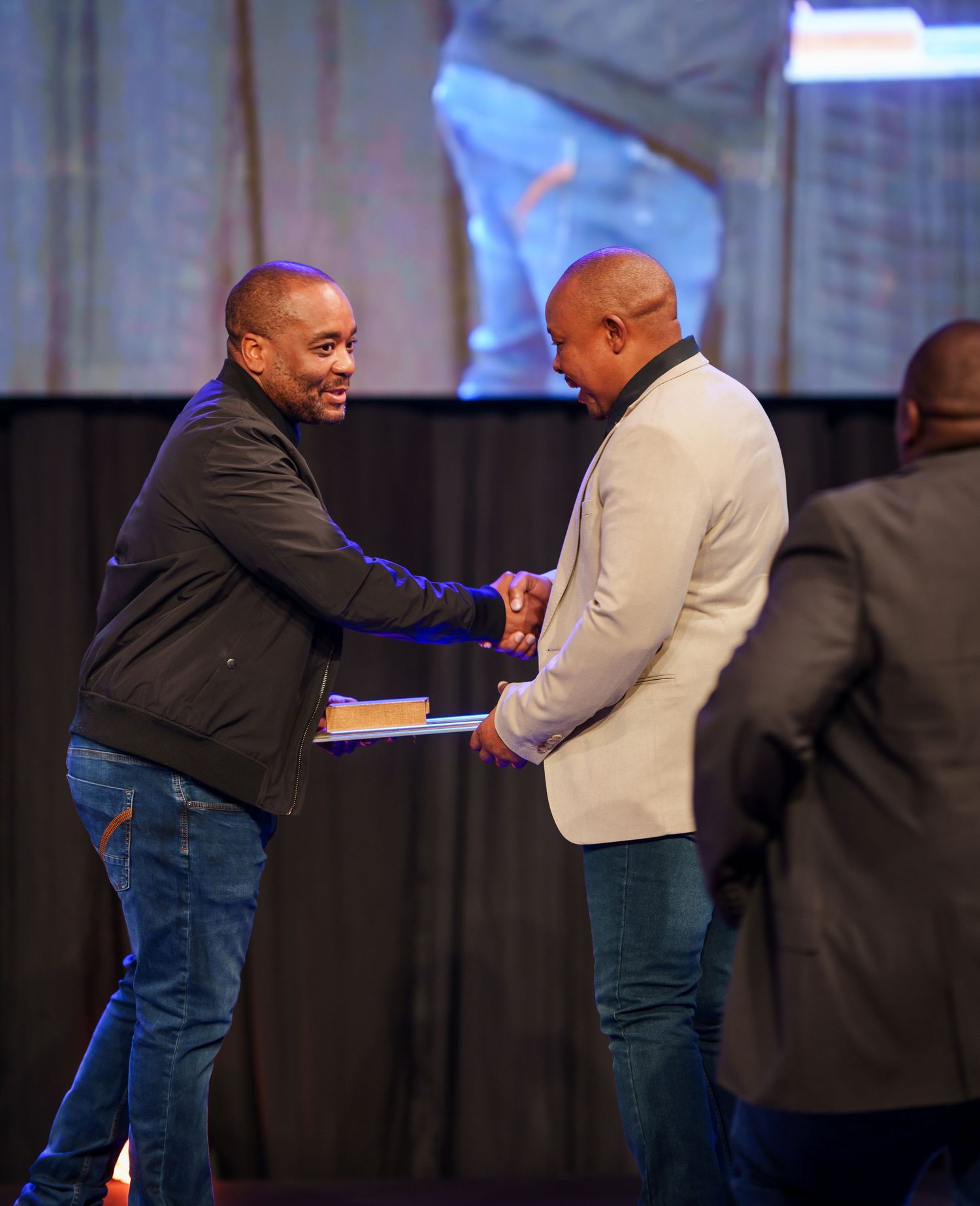 Two men exchange handshake during award presentation at formal corporate stage event.