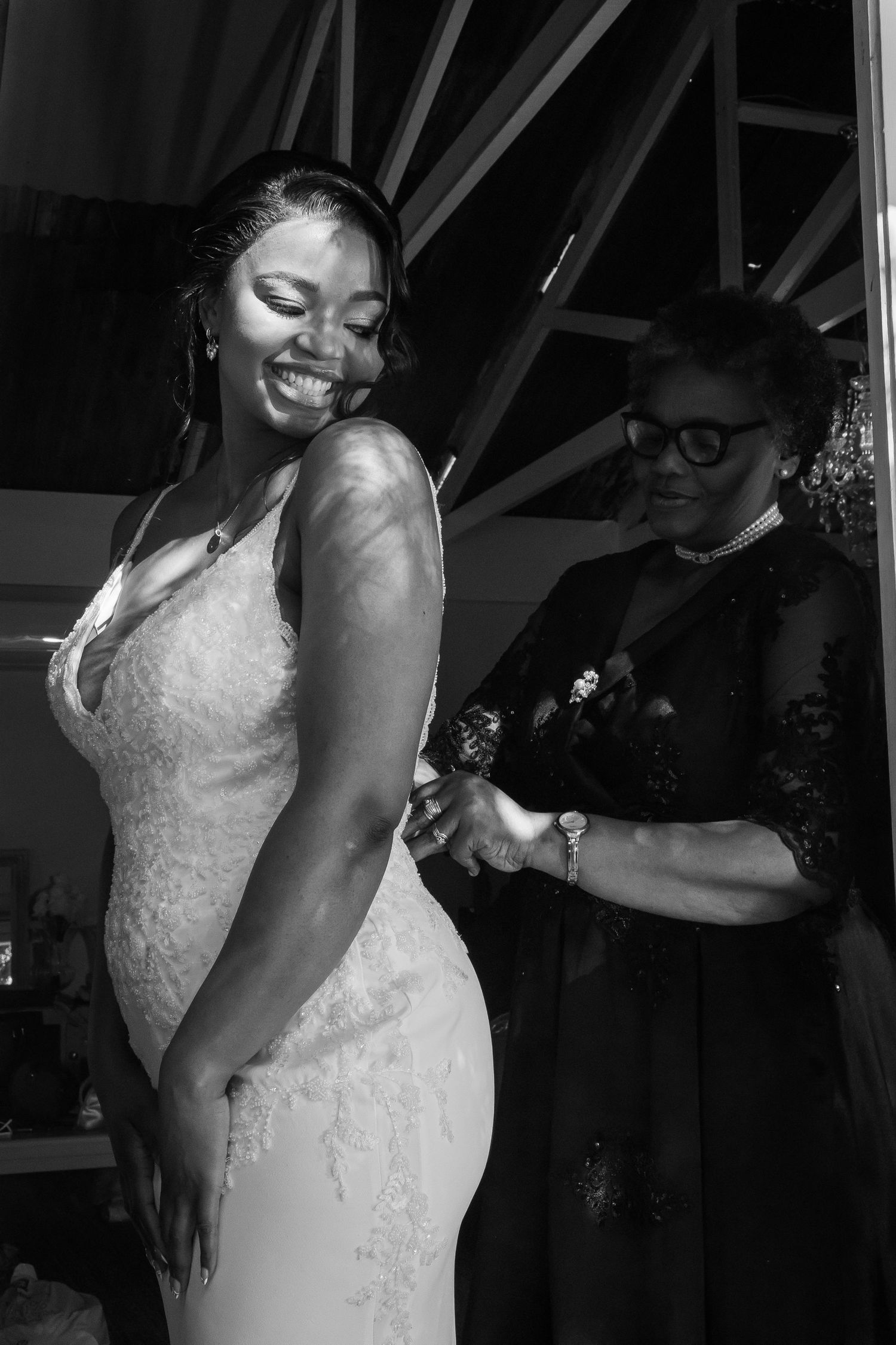Black and white photo of a smiling bride being helped into her wedding dress by a woman.
