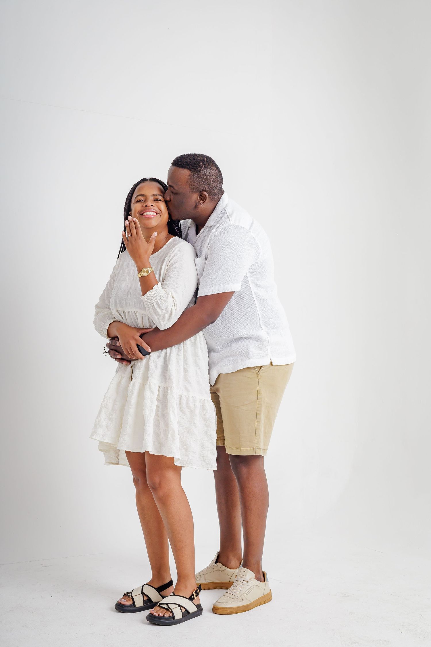 Affectionate couple in white outfits, man kissing womans cheek while embracing in white studio.