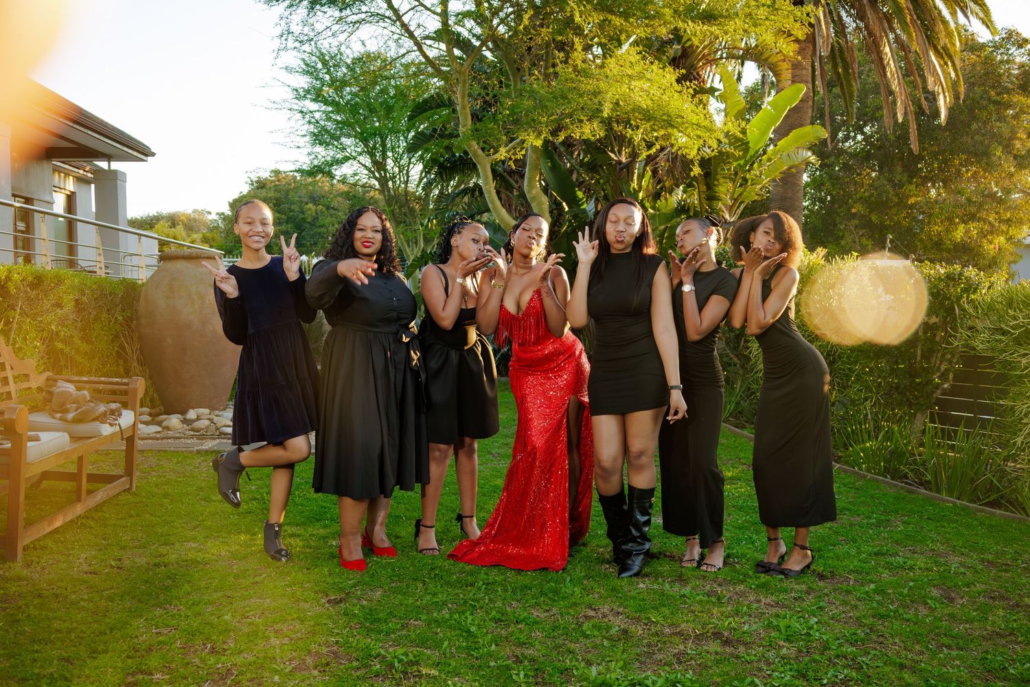 Group of women in black dresses posing outdoors with one woman in red gown, celebrating together on green lawn.