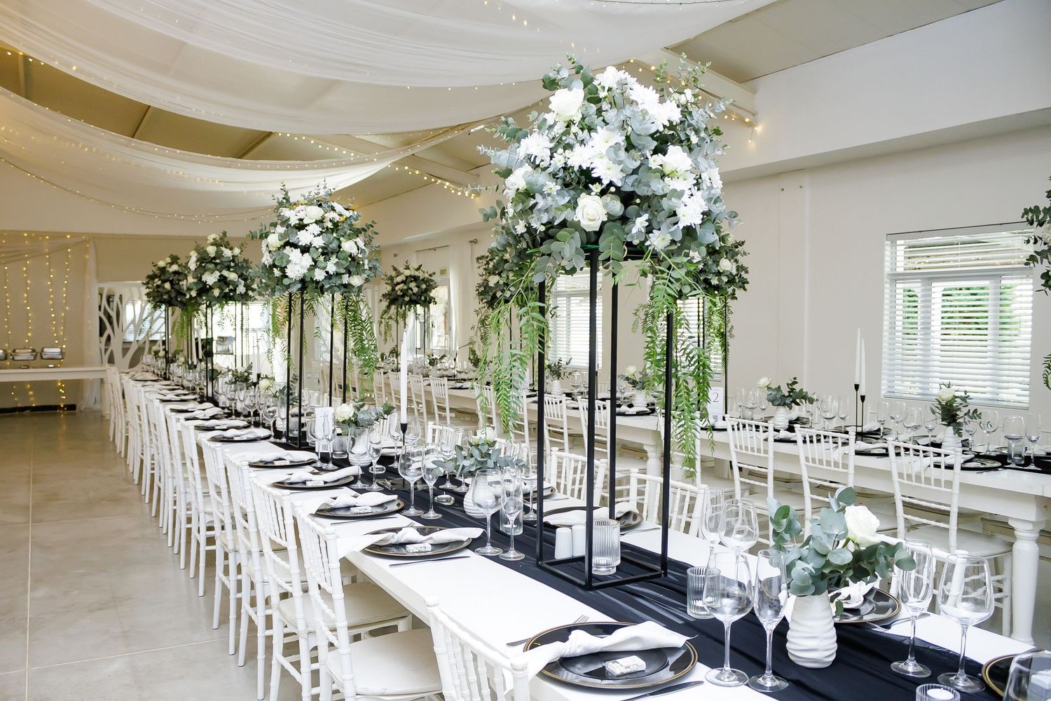 Stunning wedding reception hall with tall white floral centerpieces on long black draped tables.