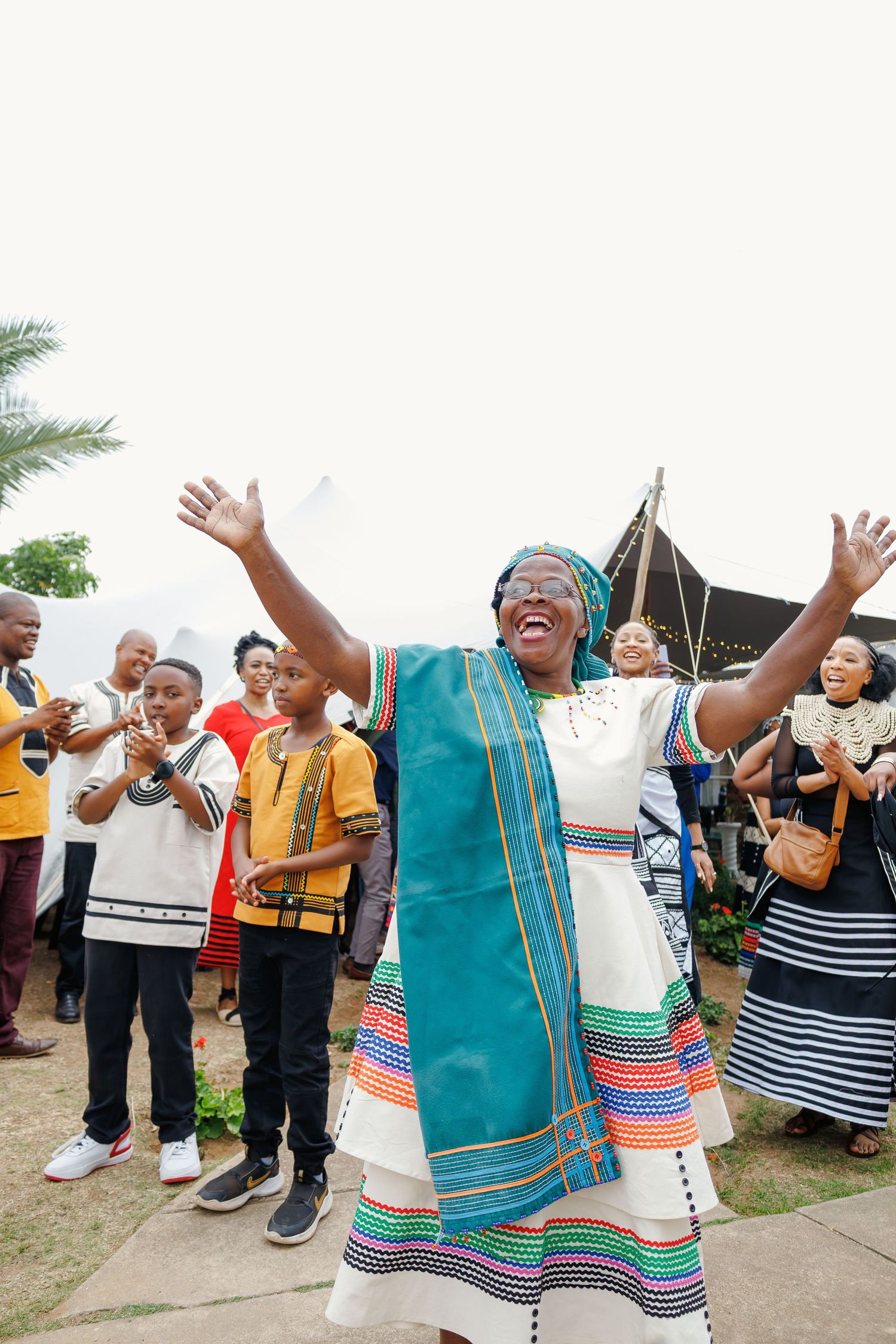 Joyful woman in colorful Xhosa traditional dress celebrates with arms raised at outdoor event.