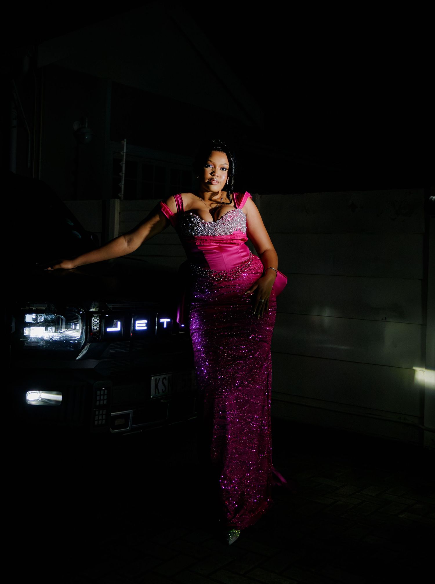Woman in sparkling pink sequin gown posing elegantly against a dark vehicle at night.
