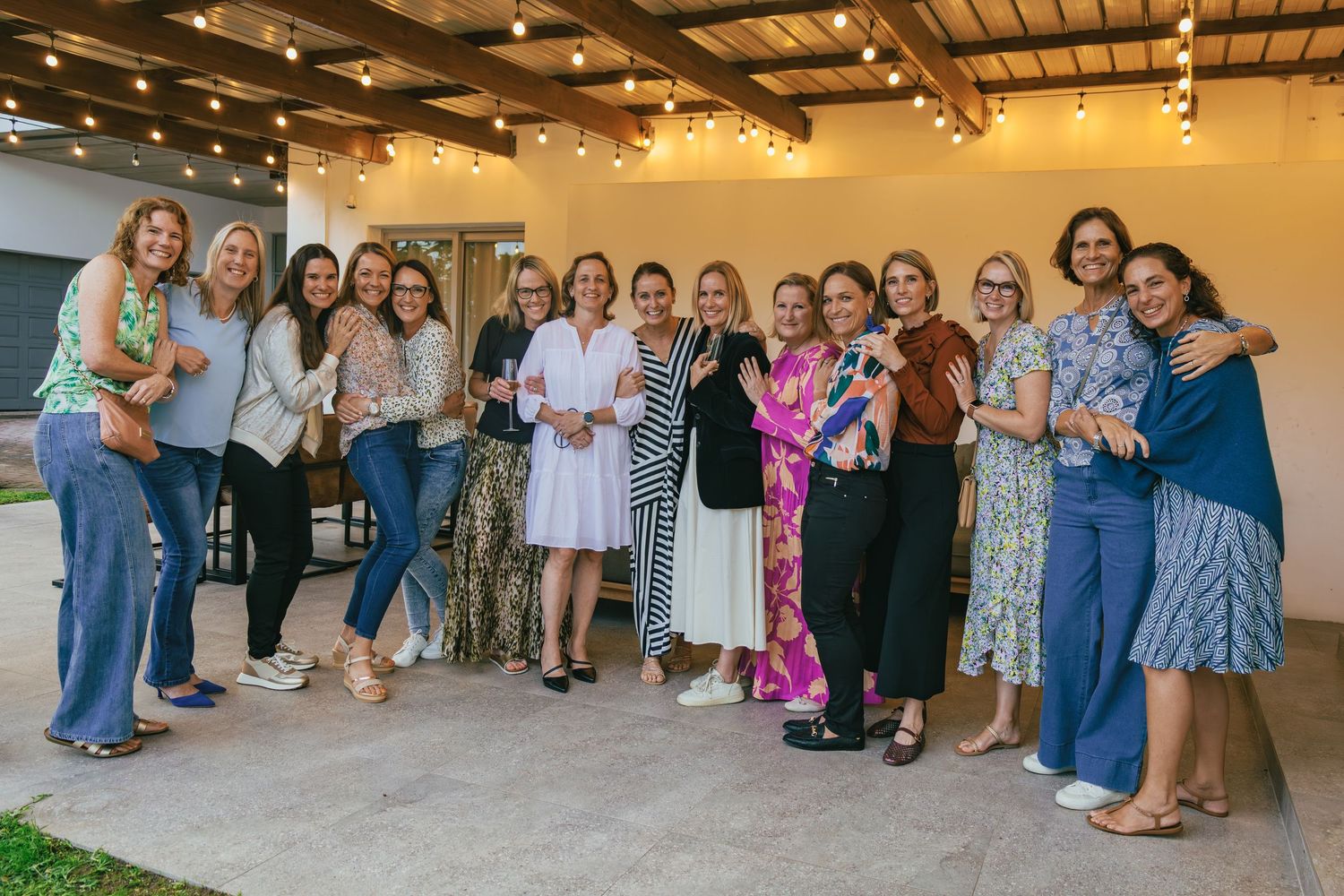 Group of women smiling and posing together at an outdoor gathering with string lights overhead.
