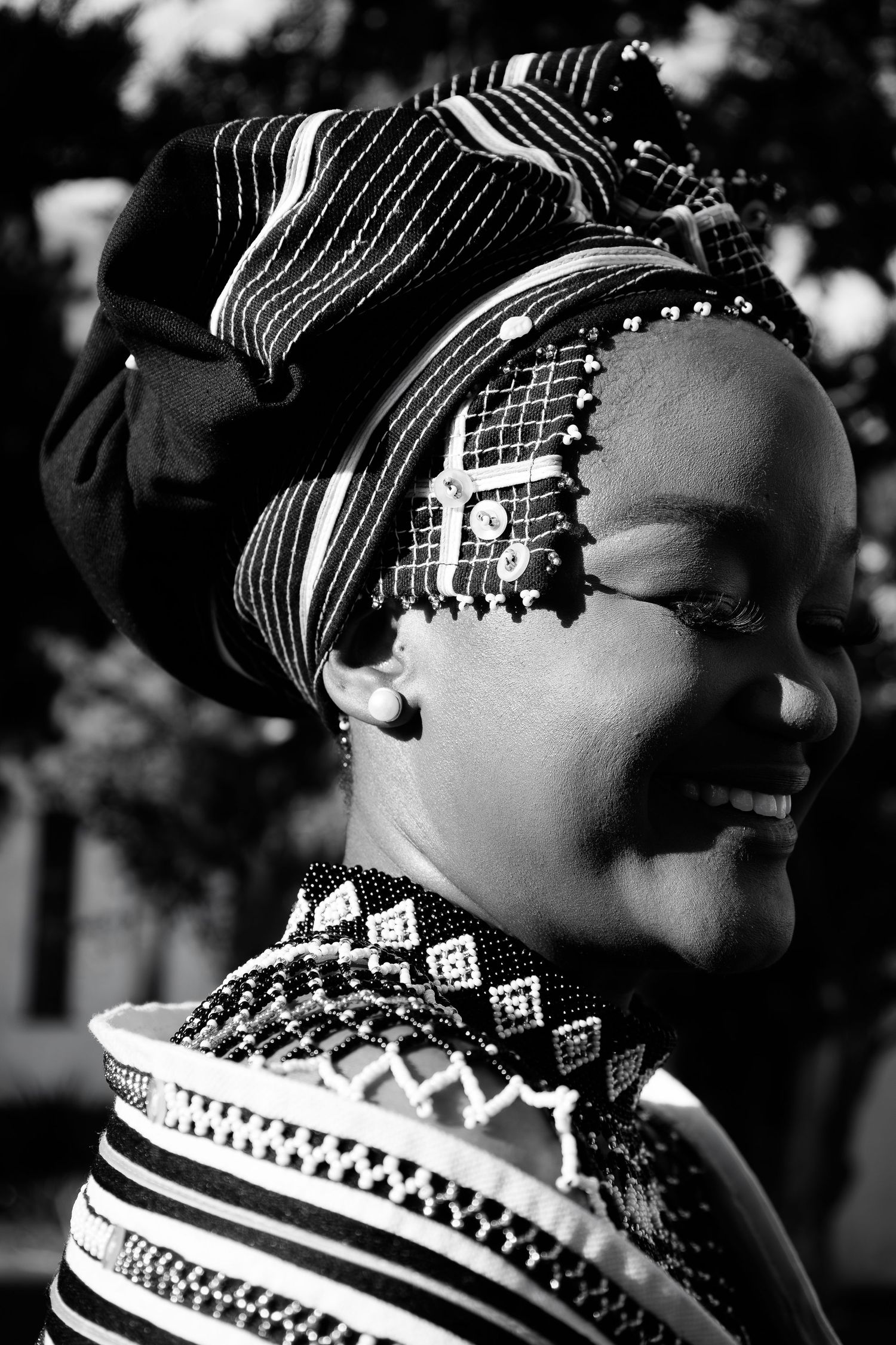 Black and white portrait of smiling woman in traditional Xhosa beaded headwear and patterned attire.