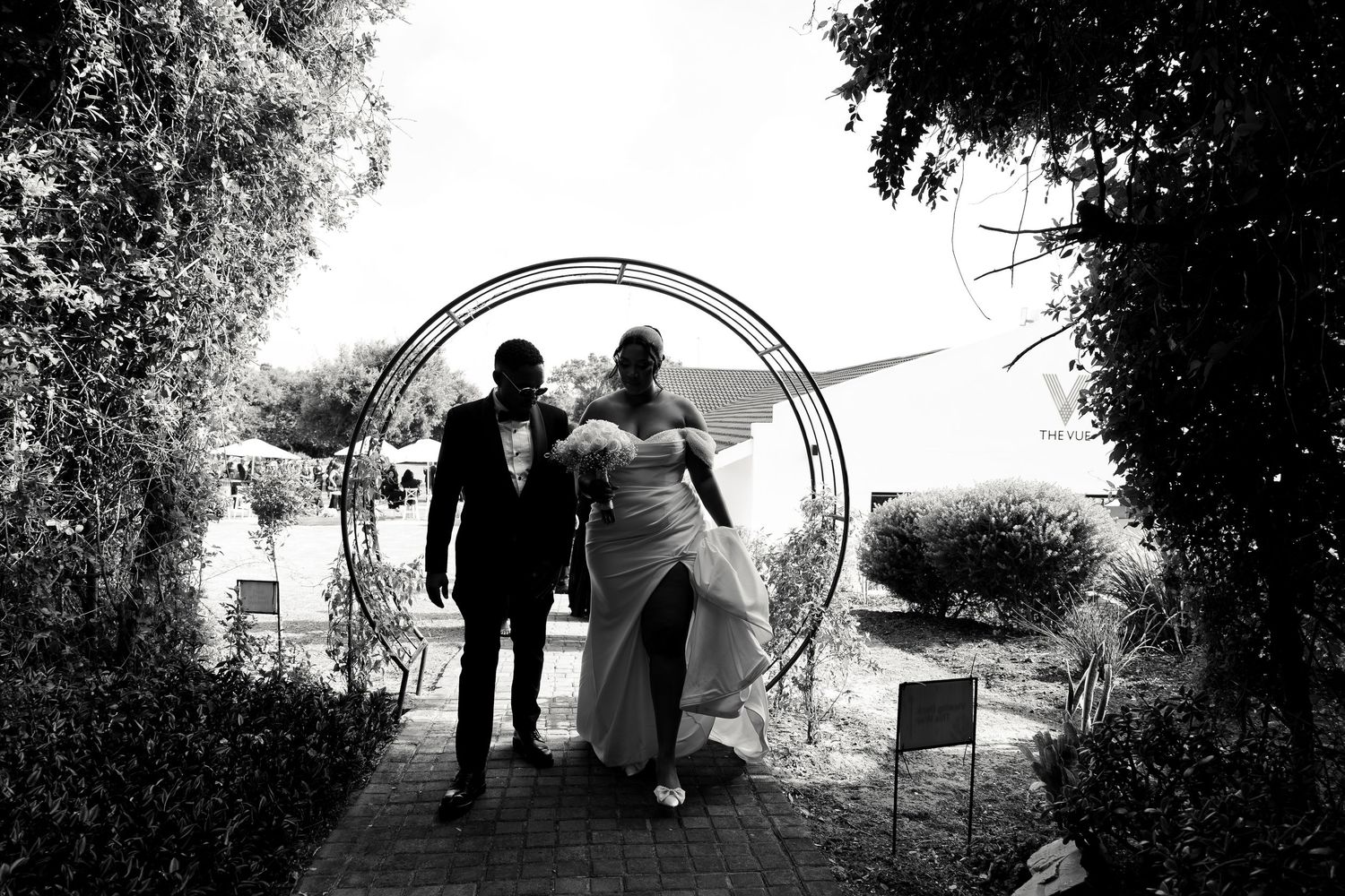 Silhouette of bride and groom walking through circular garden arch in black and white.