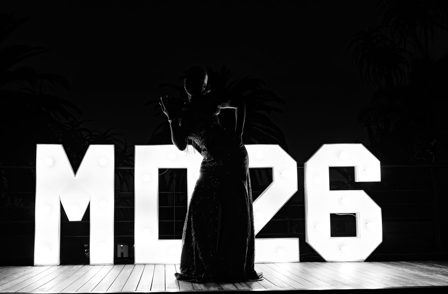 Black and white photo of a silhouetted figure posing in front of illuminated MD26 marquee letters at a celebration event.