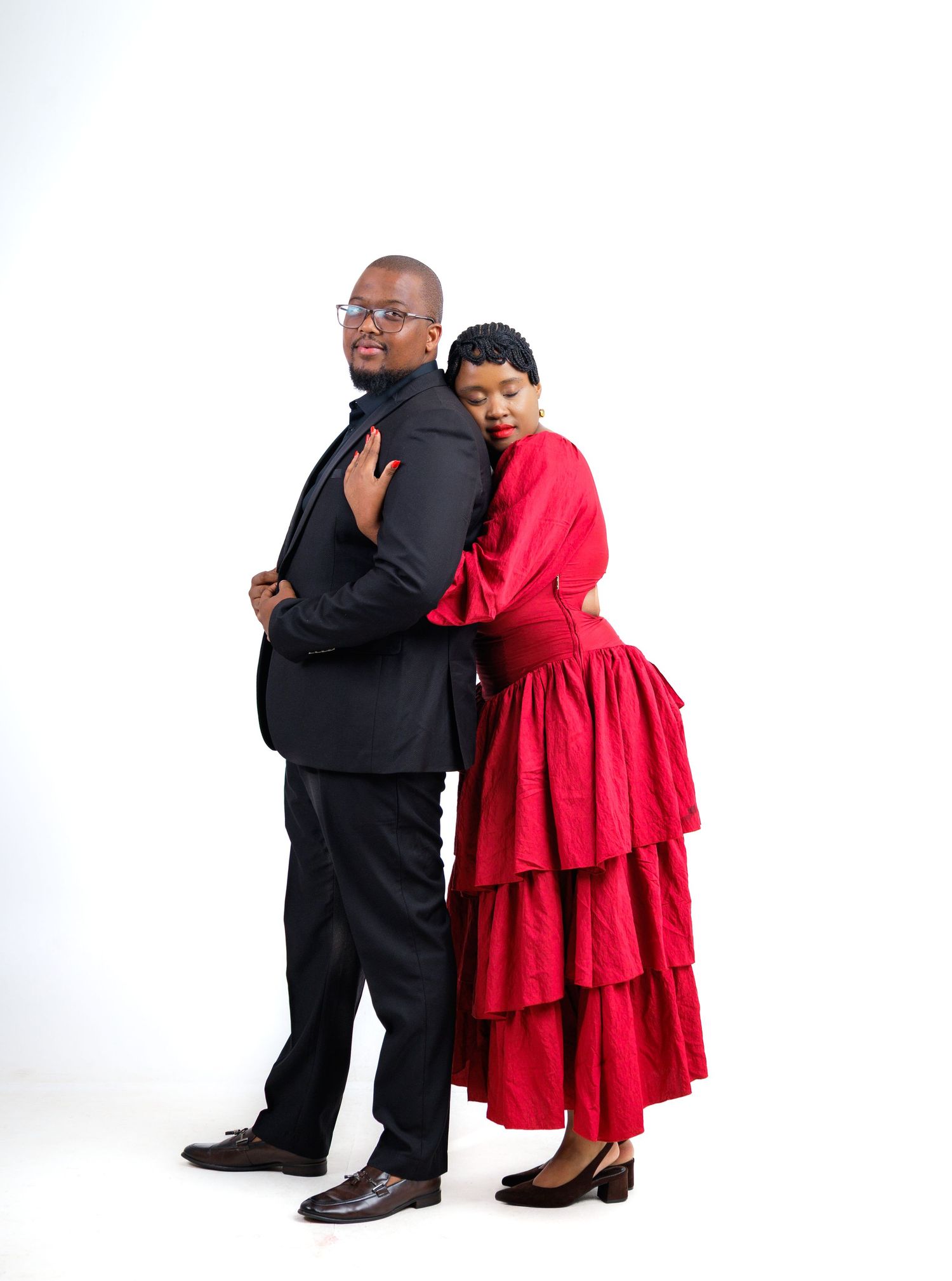 A man in a black suit and a woman in a red tiered dress pose back-to-back in a studio setting.