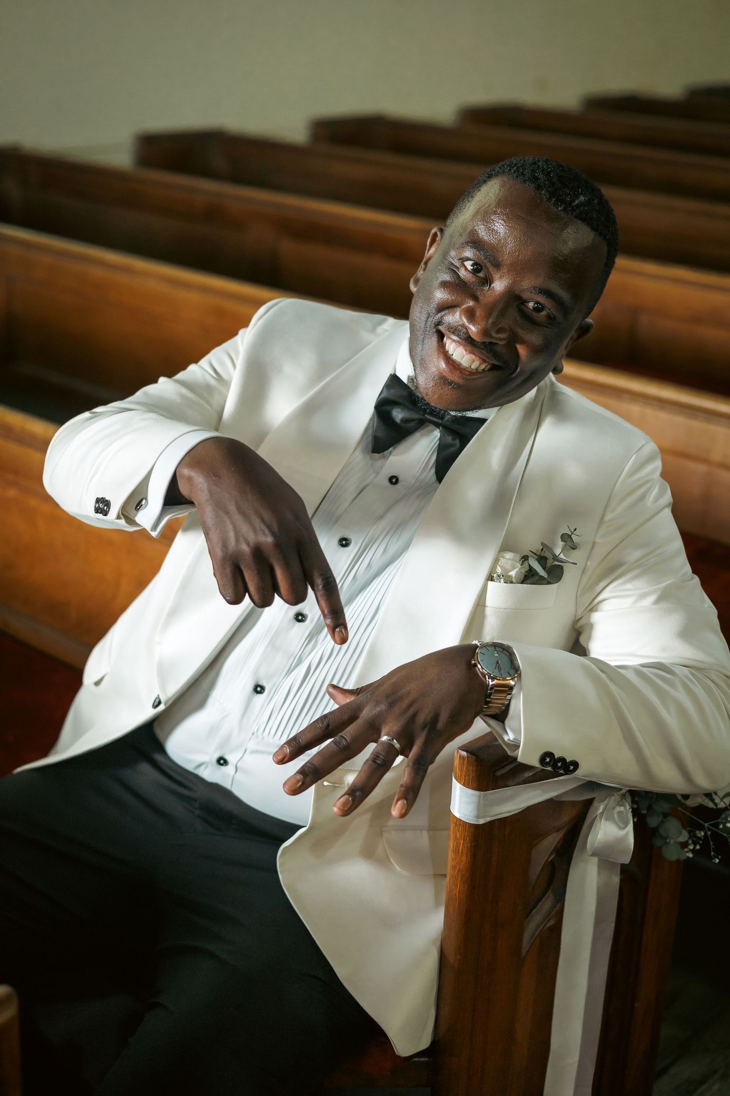 Joyful groom in a white tuxedo sitting in a church pew, smiling and pointing at his wedding ring.