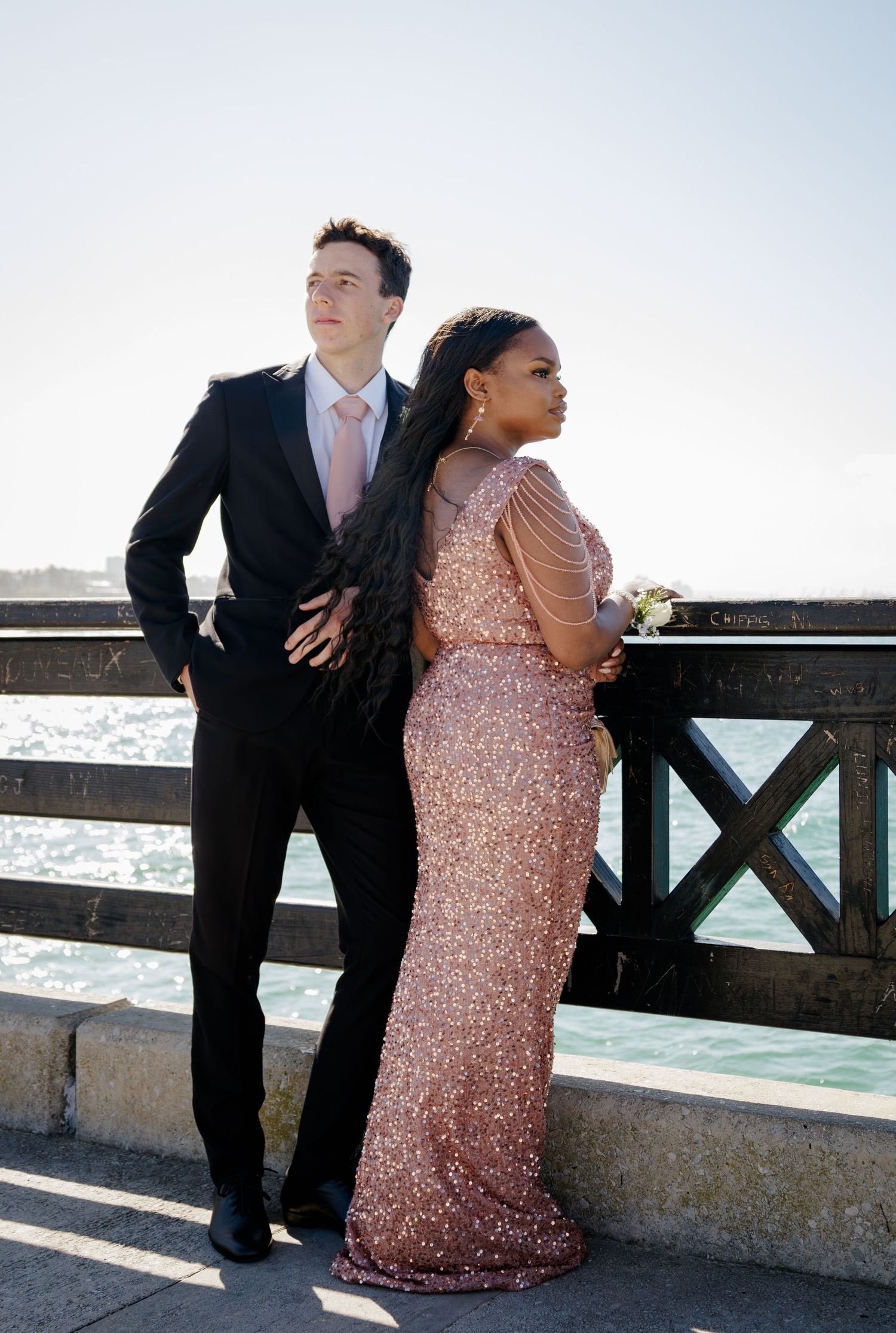 Couple in formal attire pose by waterfront railing, woman in rose gold sequin gown with corsage.