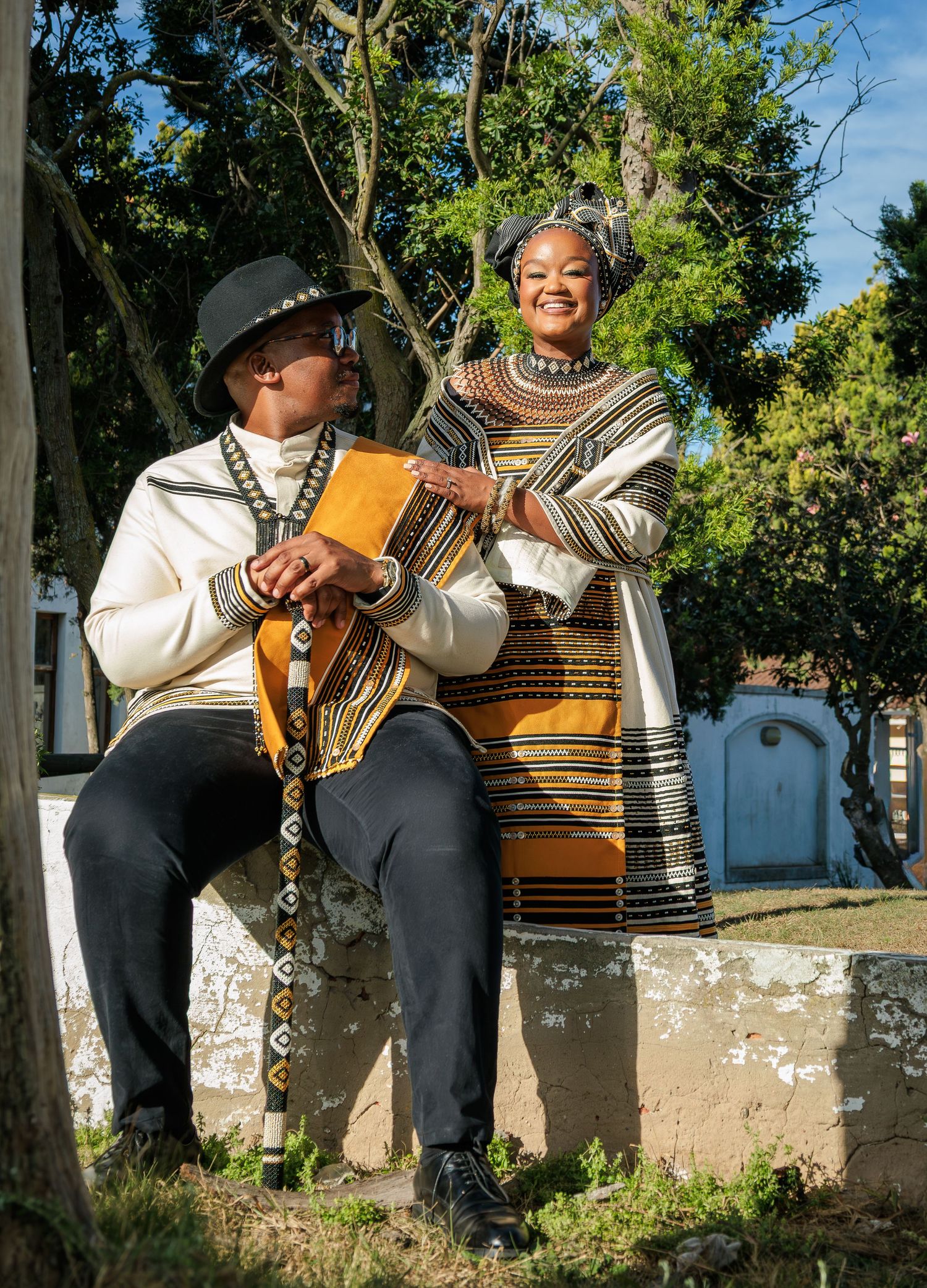 Couple posing in traditional Xhosa outfits, man seated holding stick, woman standing beside him.