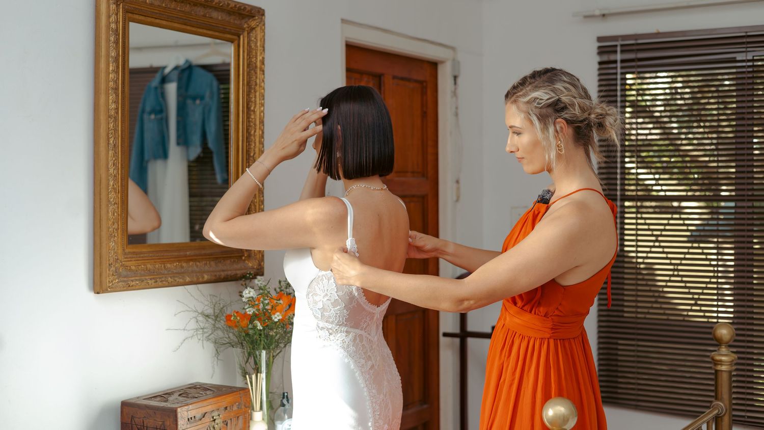 Bridesmaid in orange dress helping bride in white lace gown prepare in front of gold-framed mirror.