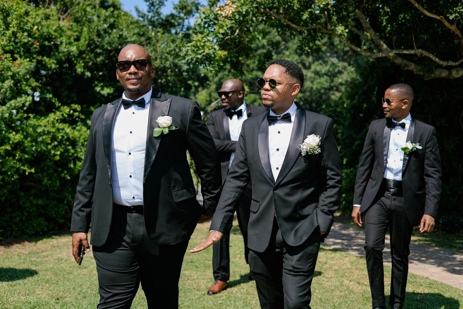 Groom and groomsmen in black tuxedos with white boutonnieres walking outdoors in bright sunlight at a wedding.